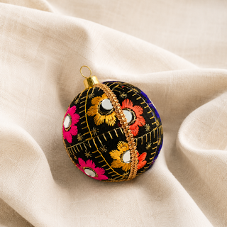 Vimal bauble hanging with soft bokeh lights, showing its black ground and bursts of gold, orange, pink and blue floral embroidery with mirror discs and gold quilted stitch lines.