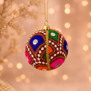 Vivaan bauble hanging with soft bokeh lights, showing its deep teal (also in maroon) ground and densely embroidered peacock-feather motifs in green, pink, orange and blue with mirror-disc centres.