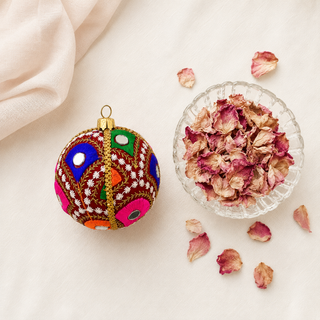 Vivaan fabric bauble styled on linen, an 8cm handmade ornament with densely embroidered peacock-feather motifs in green, pink, orange and blue with mirror-disc centres.