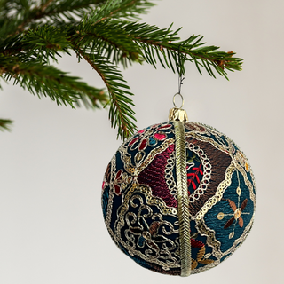 The Aaina Bauble hanging from a pine tree branch against a soft neutral background. Rich teal fabric embroidered with ornate gold, red, and blue floral motifs.