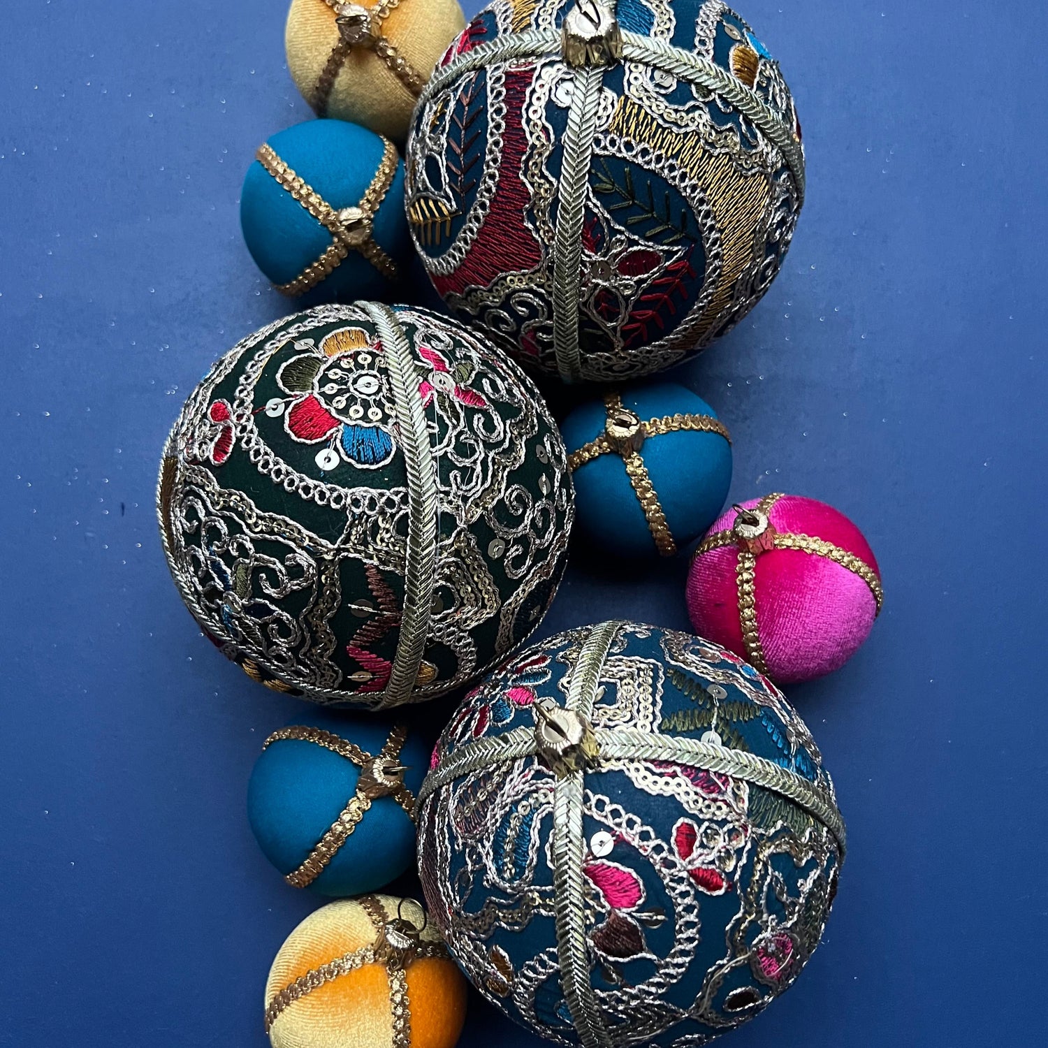 Flat lay of Aaina Baubles arranged with smaller coordinating baubles in rich jewel tones. Embroidered in gold thread with multicolour accents, styled on a deep blue background to showcase texture and colour variety.