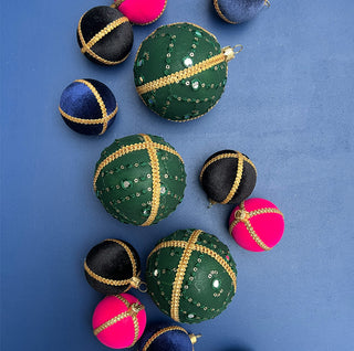 Flat lay of assorted handmade baubles on a blue background, with the Akansh Bauble in 8cm standing out in rich green and gold among colourful velvet designs.