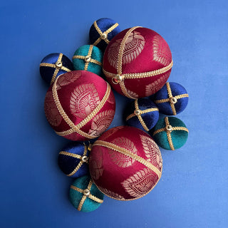 A flatlay of three Anant Baubles in deep red velvet with gold trim, styled with coordinating blue and green baubles against a vibrant blue background.