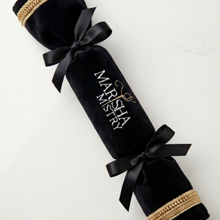 A black velvet Christmas cracker ornament with gold braided trim and black ribbon bows, featuring the personalized name 'MARISHA MYSTERY' in white and gold script, laid diagonally on a light off-white background.