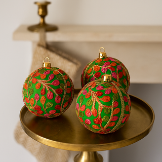 Three decorative Devaki Christmas balls with green, red, and gold patterns on a gold stand.