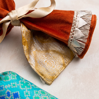 A close-up of a folded rust-orange velvet fabric item tied with a cream satin ribbon, showing a gold brocade patterned underside and a silver-and-white fringed trim. Part of a turquoise patterned textile is visible in the lower left on a light neutral background.