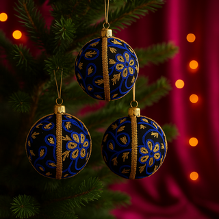 Trio of 8cm Ishana Baubles hang naturally on a green tree with a magenta velvet backdrop and gold bokeh lighting, featuring bold blue and gold floral embroidery.