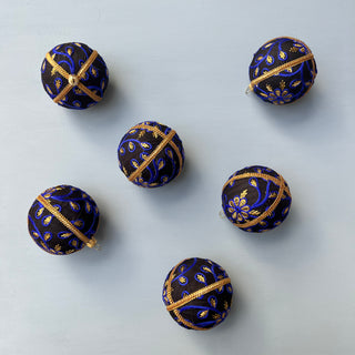 Flat lay of multiple Ishana Baubles in 8cm with black fabric, gold trim, and bright blue and gold embroidery, arranged on a soft blue background.