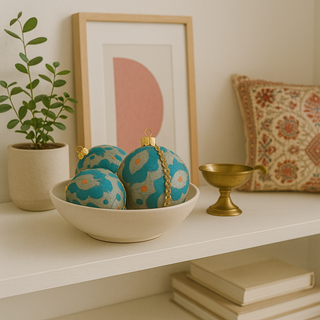 Three Johar Baubles styled in a ceramic bowl on a white shelf, surrounded by books, a plant, a brass diya, and an embroidered cushion.