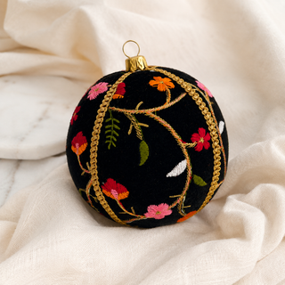 A round black Christmas ornament decorated with colorful floral embroidery and gold braided trim, resting on draped cream fabric.