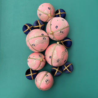 A group of Lalika Baubles in 8cm, 10cm, and mini sizes laid flat on a teal background, showcasing pink velvet, gold trim, and purple floral brocade embroidery in a luxurious, colour-rich setting.