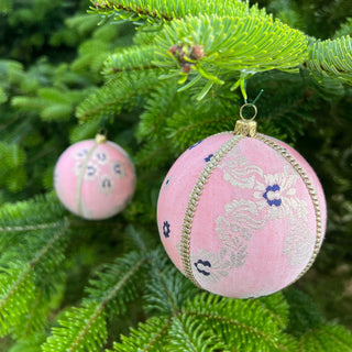 Two Lalika Baubles made of pink velvet with gold trim and purple floral brocade hanging from the branches of a real Christmas tree outdoors.