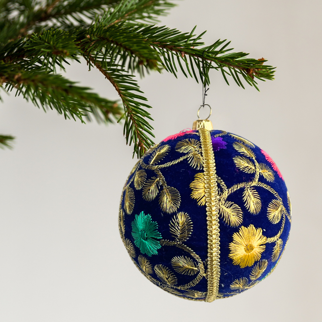 Decorative Mihir Christmas ball ornament with floral patterns on a tree branch. Handcrafted Mahir Bauble in navy velvet with intricate gold thread and floral embroidery, UK-made.