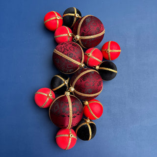 A cluster of decorative Christmas ornaments shaped like small balls, featuring a mix of glossy red, deep burgundy damask-patterned, and matte black ornaments, each detailed with gold metallic trim, set against a solid blue background.