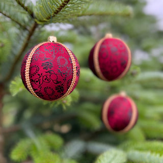 Three round red Christmas ornaments with black floral patterns and gold trim hanging on green evergreen tree branches.