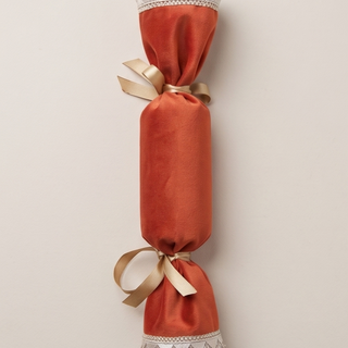 An orange fabric Christmas cracker ornament with gold ribbon bows and silver lace trim, shown vertically against a light beige background.
