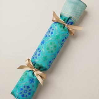 A cylindrical party cracker or decorative paper roll wrapped in turquoise and blue patterned fabric, tied with two beige ribbon bows, photographed on a plain light beige background.