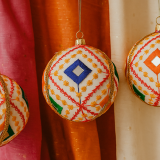 Three Sohan Baubles hanging against colourful draped fabric, showing vibrant mirrorwork embroidery in red, orange, green and blue on a cream base.