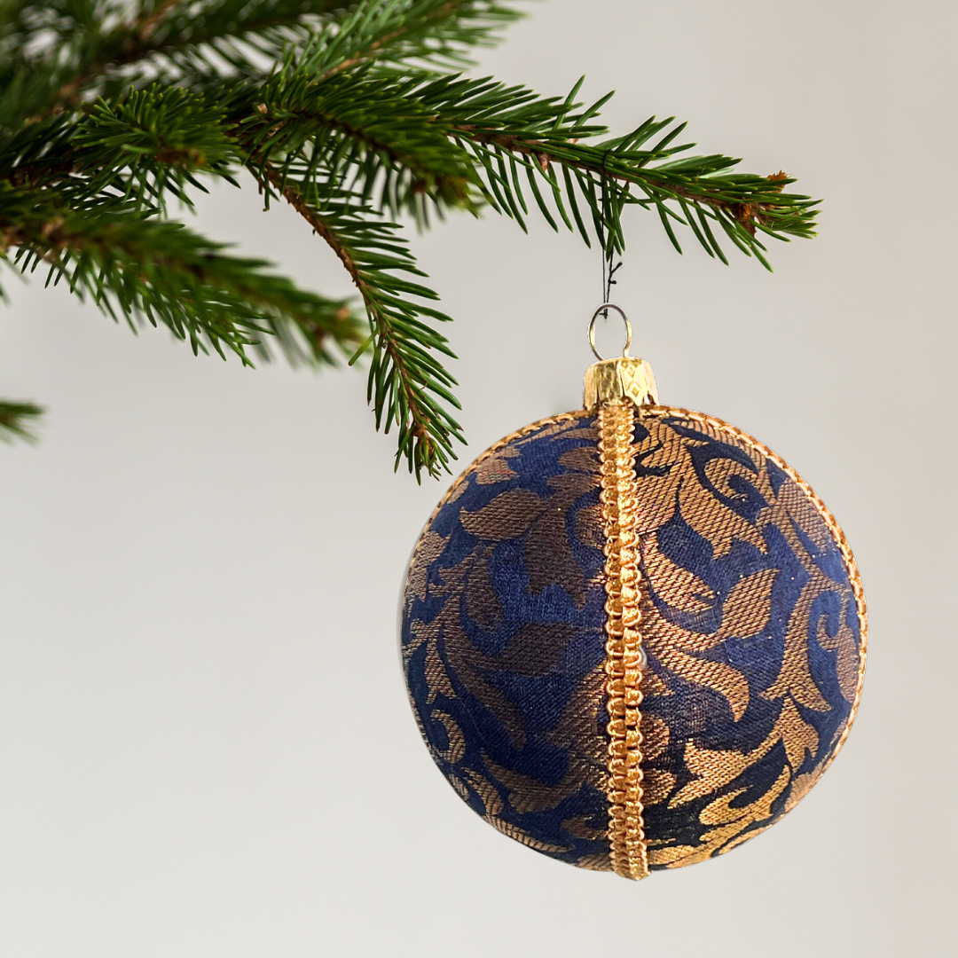 Decorative Christmas ball ornament with gold and blue pattern hanging on a tree branch. Vani Bauble – sustainable fabric decoration with baroque detailing, handmade in the UK.