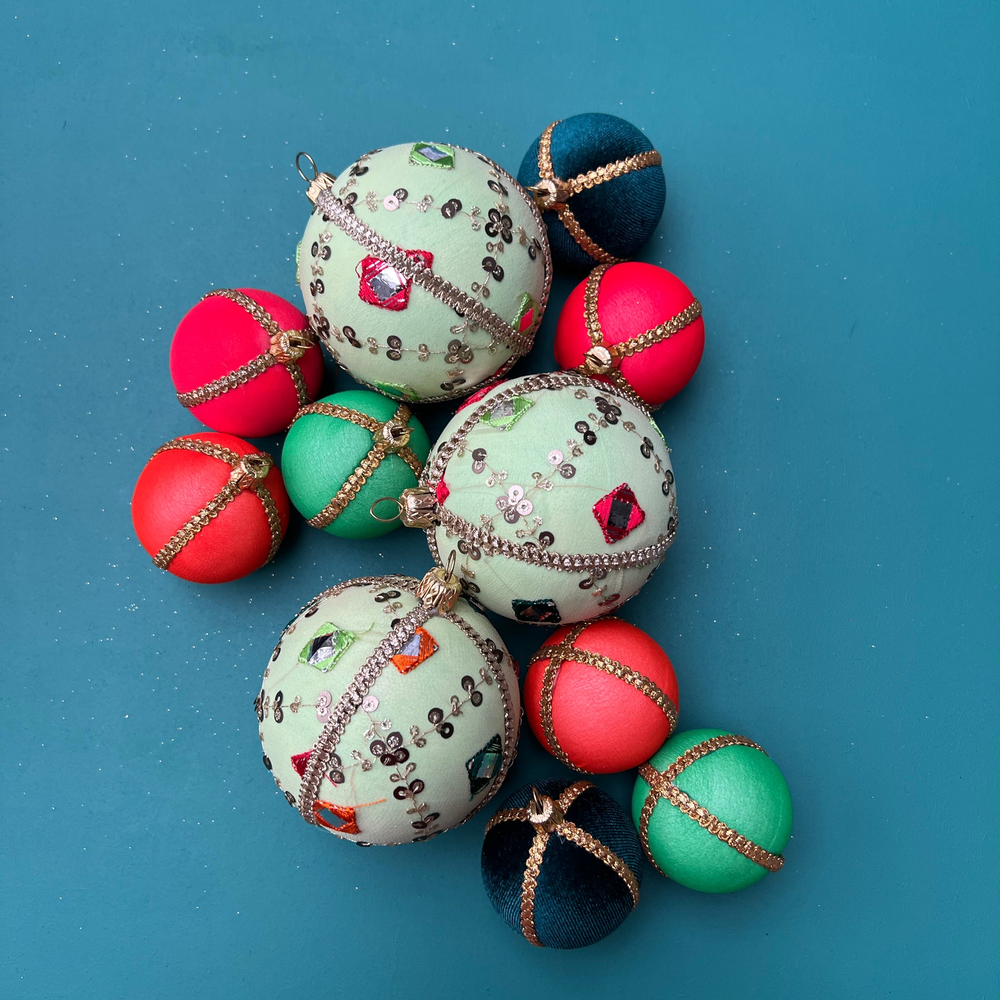 
"Three light green velvet Gauri baubles with colourful mirror embroidery and gold trim, styled alongside contrasting mini baubles in rich jewel tones, laid flat on a bold teal background.