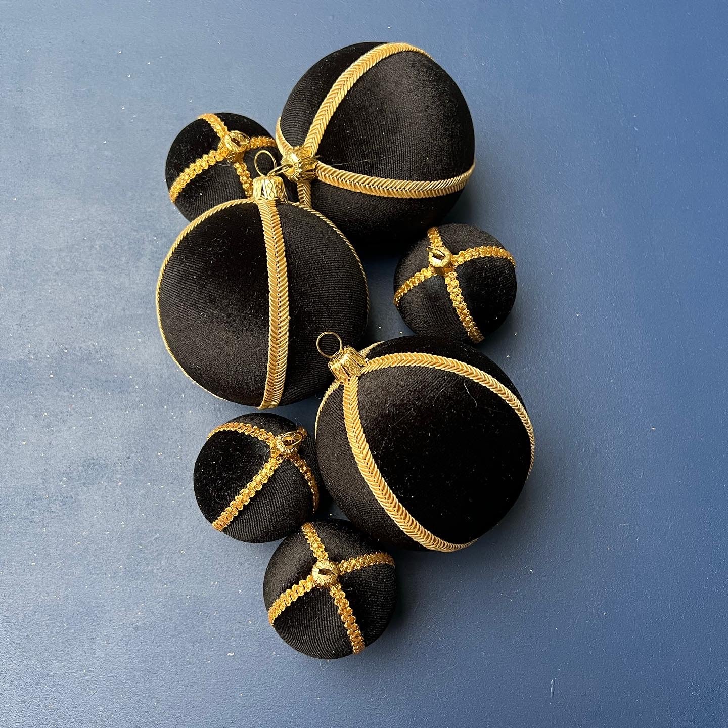 Four black velvet Christmas baubles with gold trim arranged flat on a bold blue background, showcasing their luxurious texture and metallic details in a modern, minimal setting