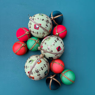Three light green velvet Gauri baubles with colourful mirror embroidery and gold trim, styled alongside contrasting mini baubles in rich jewel tones, laid flat on a bold teal background.